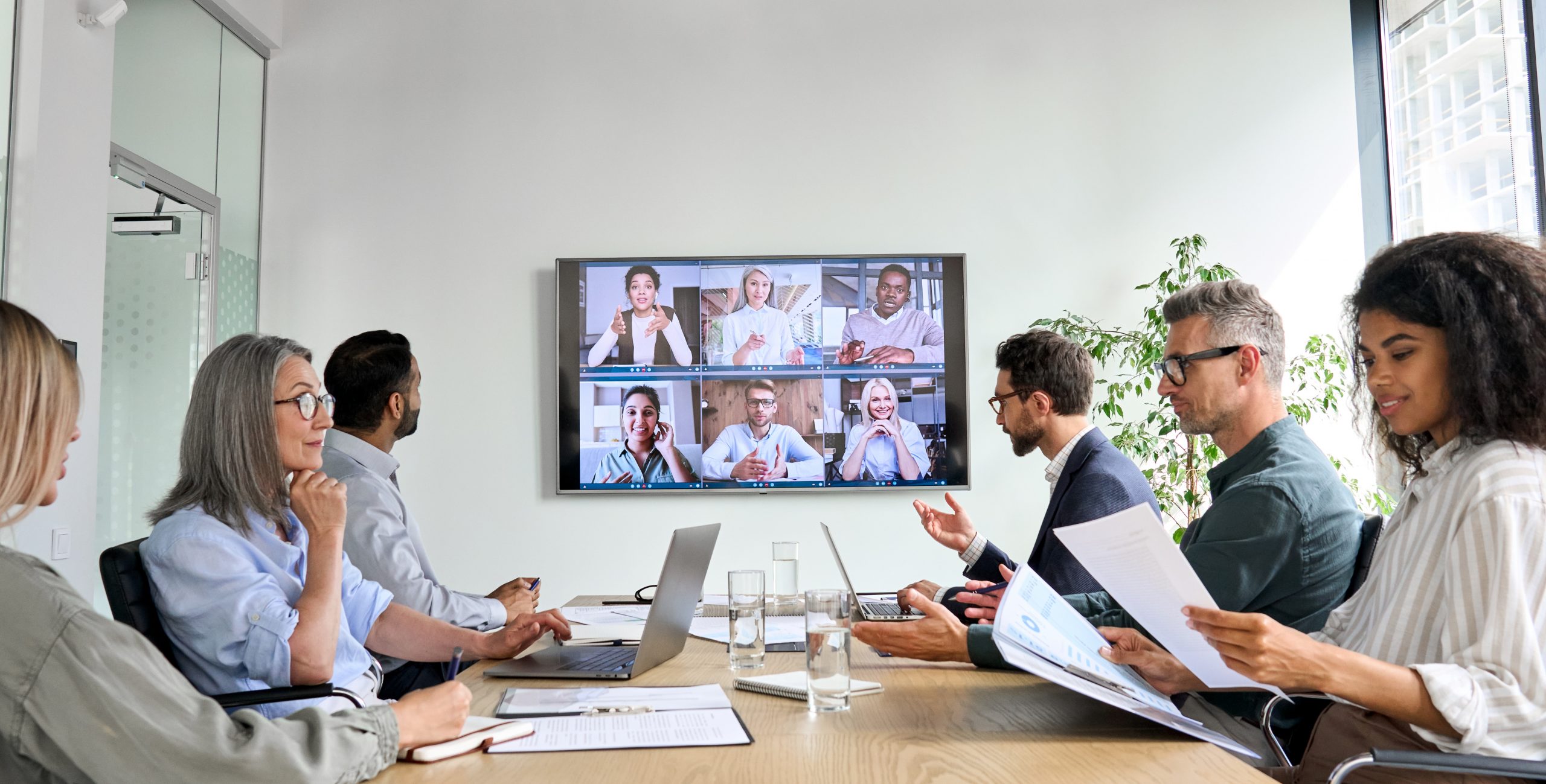 Diverse employees on online conference video call on tv screen in meeting room. Six people sitting around conference table having meeting with six other people via tele-conferencing call